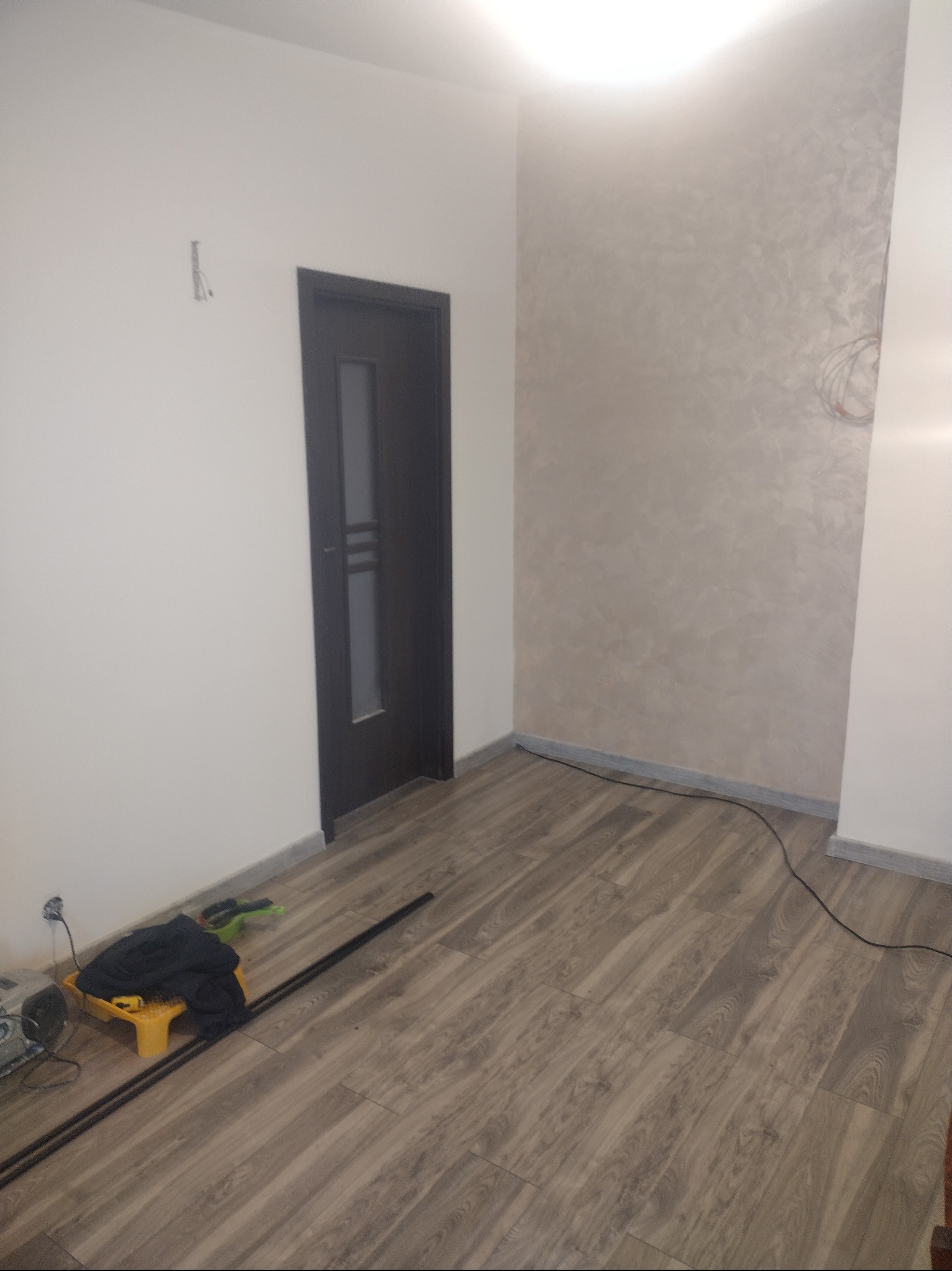 Wnętrze pokoju w trakcie prac wykończeniowych: położona podłoga z paneli, ściana z dekoracyjną strukturą, zamontowane drzwi wewnętrzne, widoczne narzędzia i akcesoria malarskie.