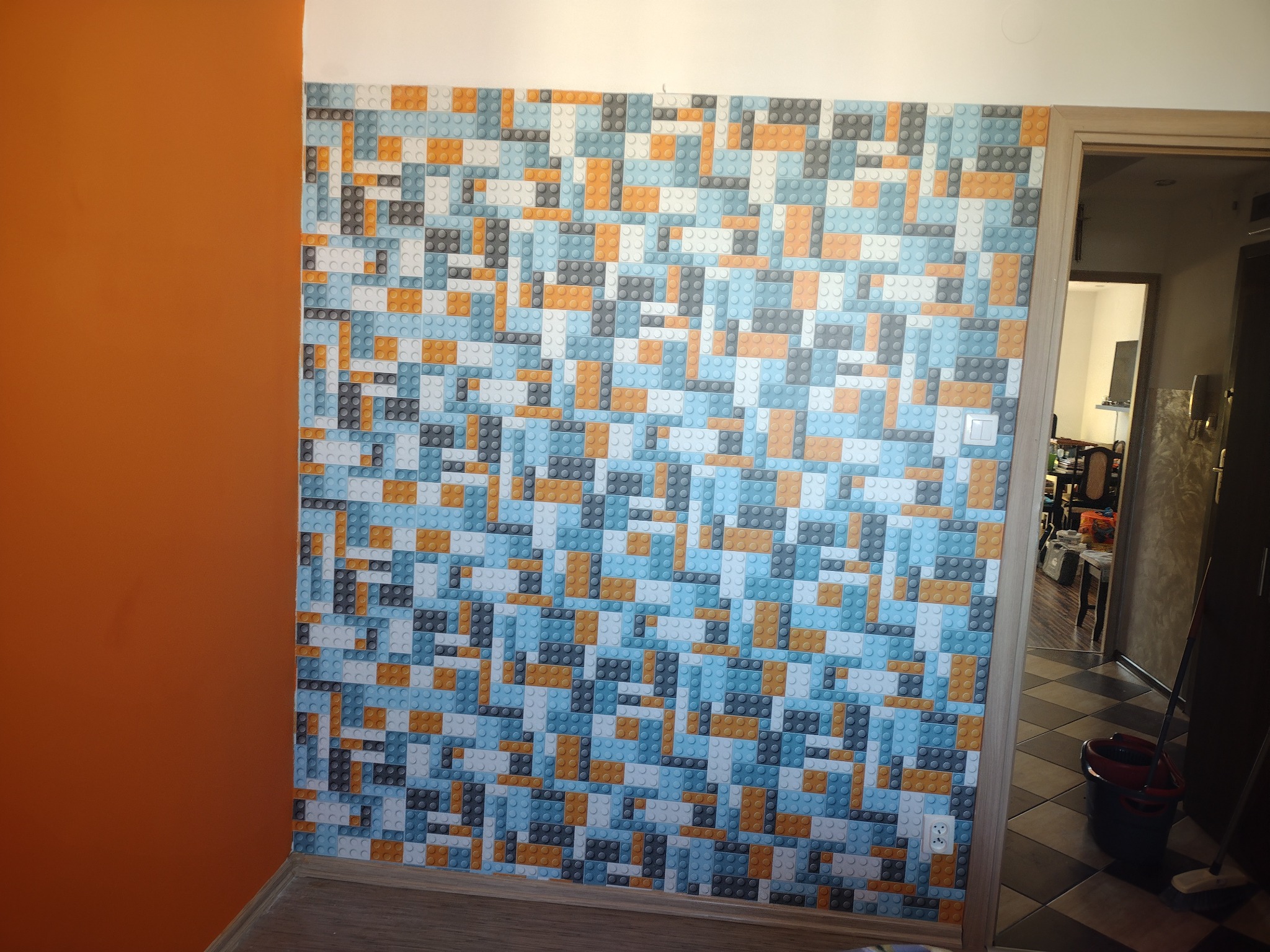 Ściana wykończona tapetą z motywem klocków Lego w odcieniach niebieskiego, szarego, pomarańczowego i białego, sąsiadująca z pomarańczową ścianą. W tle widoczne otwarte drzwi do kolejnego...