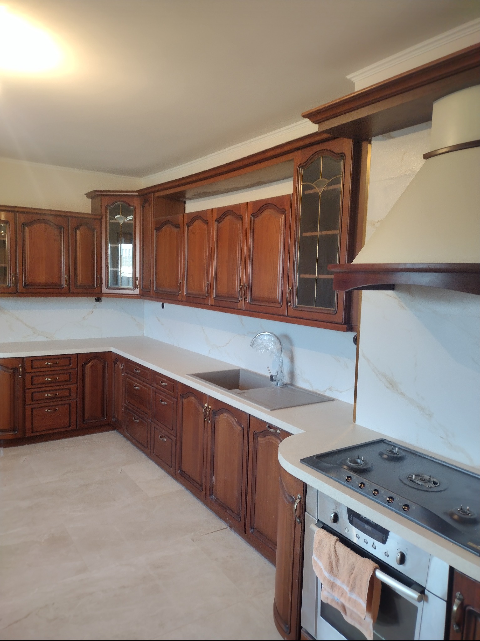 Klasyczna kuchnia z drewnianymi szafkami w ciepłym odcieniu, jasnym blatem imitującym marmur i beżową podłogą z płytek, widoczny okap i płyta grzewcza.