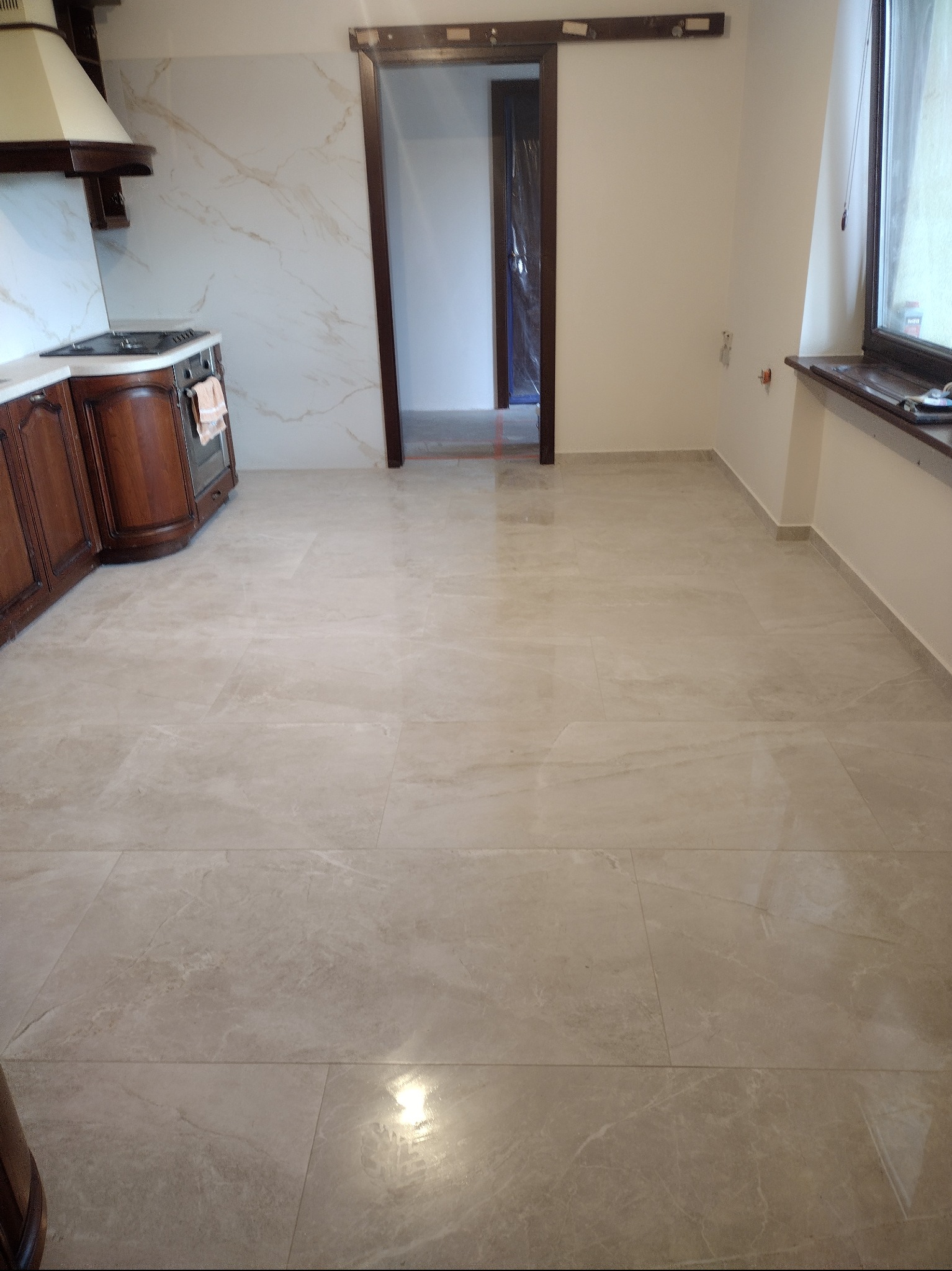 Wykończona podłoga z płytek imitujących marmur w jasnej tonacji, widok na otwarte przejście do innego pomieszczenia i fragment zabudowy kuchennej z drewnianymi szafkami.