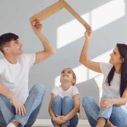 kredytylublin.pl - Kredyty, pożyczki, ubezpieczenia - Uśmiechnięta rodzina siedzi na podłodze, trzymając kartonową ramę w kształcie dachu nad głową dziecka, symbolizując bezpieczny dom.