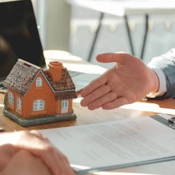 kredytylublin.pl - Kredyty, pożyczki, ubezpieczenia - Makieta domu jednorodzinnego stoi na biurku obok dokumentu, nad którym wisi dłoń w geście powitania lub akceptacji, w tle widoczny laptop.