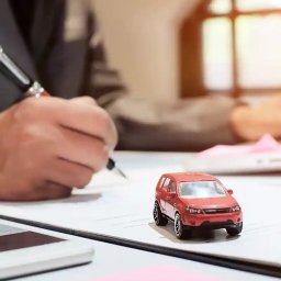 kredytylublin.pl - Kredyty, pożyczki, ubezpieczenia - Osoba podpisuje dokument długopisem, obok leży model czerwonego SUVa, tablet i różowa kartka samoprzylepna, w tle widoczne okno.