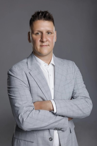 Portret mężczyzny w jasnoszarym garniturze w kratę i białej koszuli, stojącego na szarym tle z założonymi rękami.