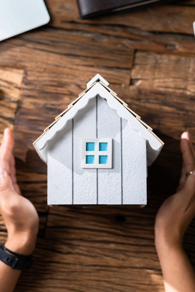 Model białego domu z niebieskim oknem na drewnianym stole, otoczony dłońmi, z widocznym zegarkiem na nadgarstku i portfelem w tle.