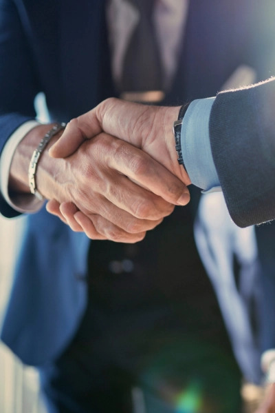 Uścisk dłoni dwóch mężczyzn w garniturach, z widocznym zegarkiem na nadgarstku i bransoletką, symbolizujący zawarcie umowy lub porozumienia.