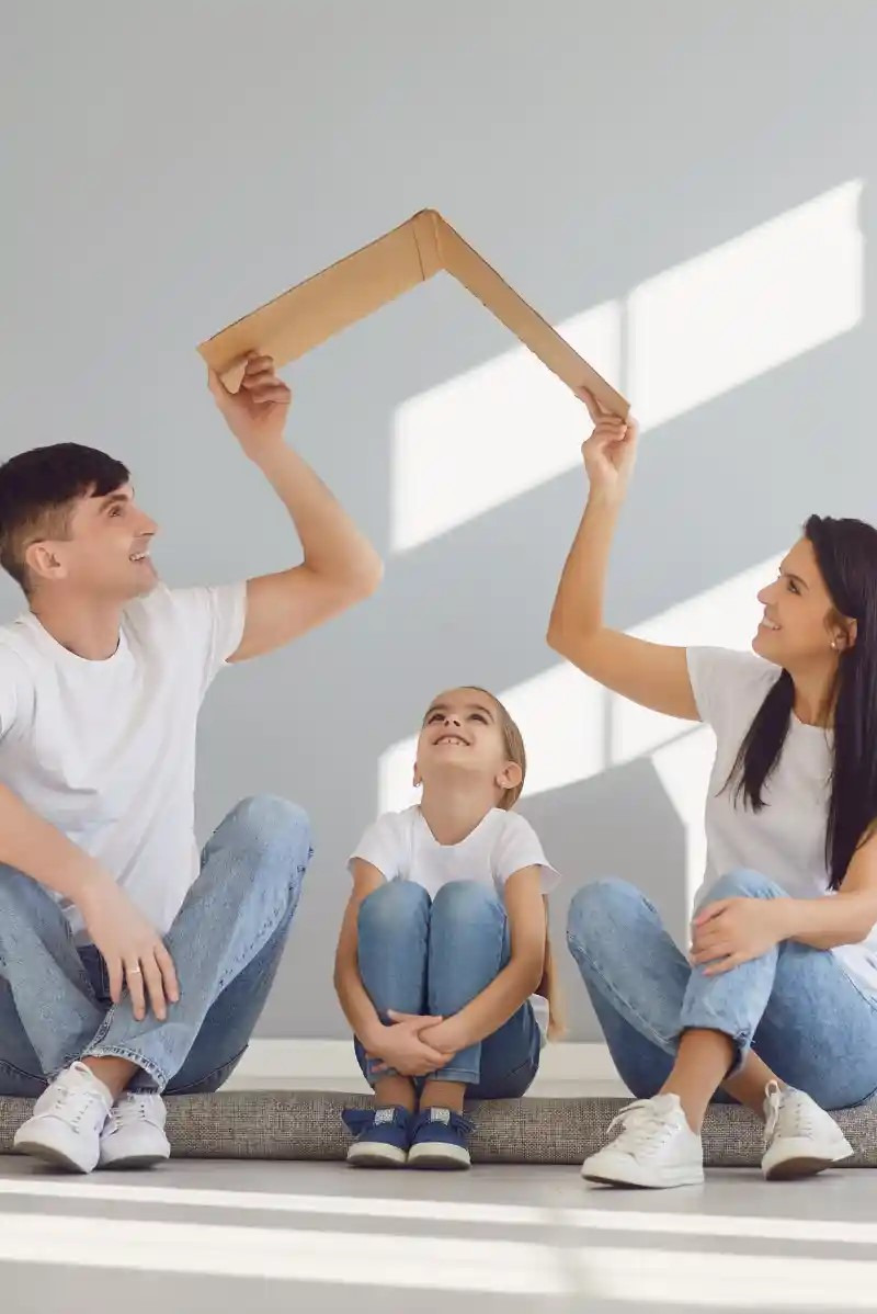 Uśmiechnięta rodzina siedzi na podłodze, trzymając kartonową ramę w kształcie dachu nad głową dziecka, symbolizując bezpieczny dom.