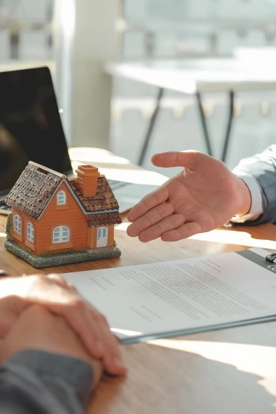 Makieta domu jednorodzinnego stoi na biurku obok dokumentu, nad którym wisi dłoń w geście powitania lub akceptacji, w tle widoczny laptop.