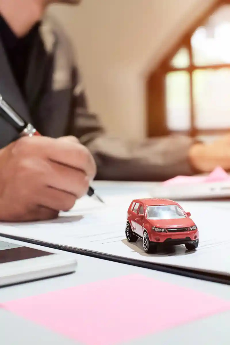 Osoba podpisuje dokument długopisem, obok leży model czerwonego SUVa, tablet i różowa kartka samoprzylepna, w tle widoczne okno.