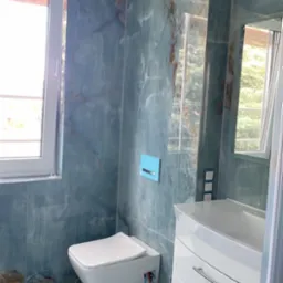 Nowoczesna łazienka z niebieskimi płytkami imitującymi marmur, podwieszana toaleta z niebieskim przyciskiem spłukującym, biała szafka pod umywalką z trzema szufladami i lustro odbijające widok z okna.