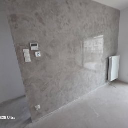 Jakość Projekt Bud Anatolii Nechyporuk - Szara ściana z tynkiem dekoracyjnym o nieregularnej fakturze, z białymi włącznikami i termostatem. Obok biały grzejnik i surowa posadzka.