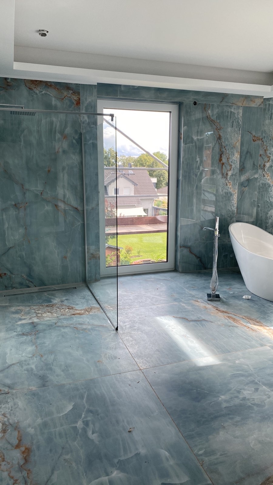Wykończona łazienka z zielonymi płytkami imitującymi marmur, kabina prysznicowa typu walk-in z przezroczystego szkła, wolnostojąca wanna i widok na zewnątrz przez okno.