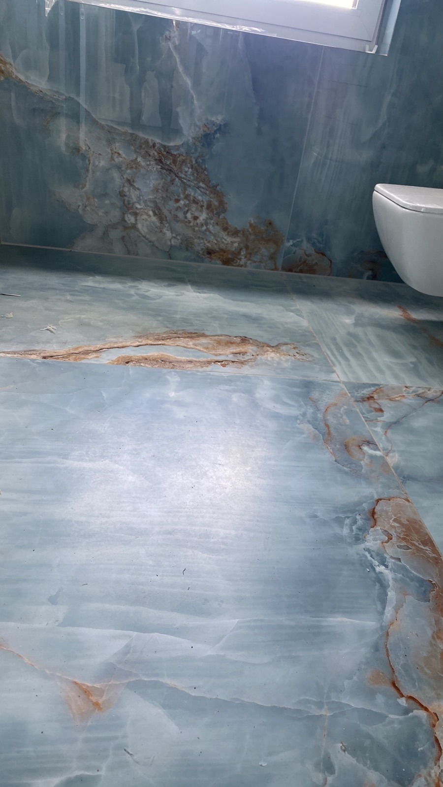 Łazienka wykończona płytkami imitującymi marmur w odcieniach błękitu i brązu, widoczny fragment toalety i okna.