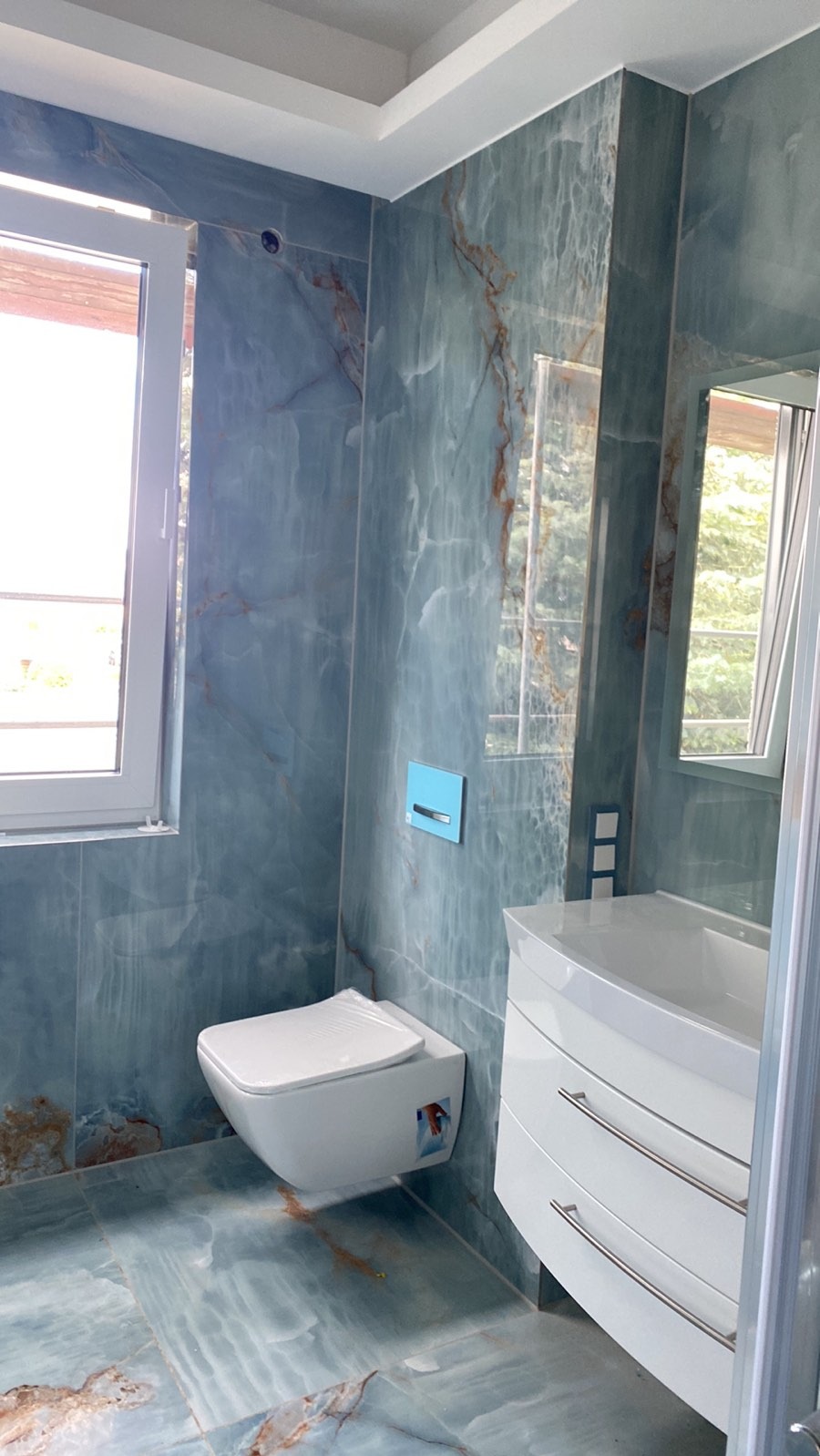 Nowoczesna łazienka z niebieskimi płytkami imitującymi marmur, podwieszana toaleta z niebieskim przyciskiem spłukującym, biała szafka pod umywalką z trzema szufladami i lustro odbijające widok z okna.