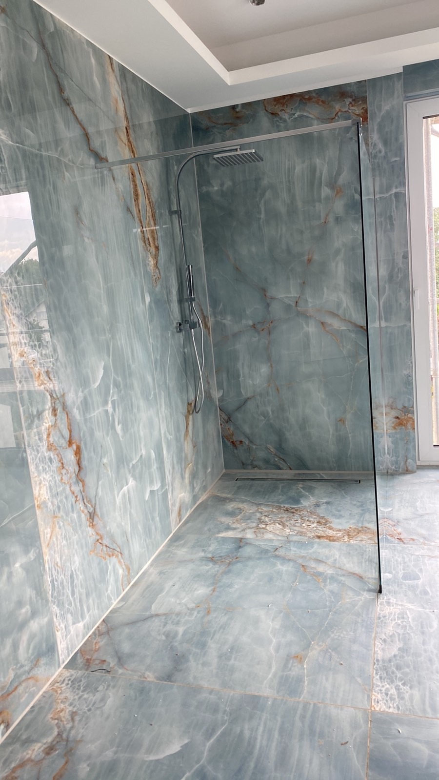 Wykończona łazienka z kabiną prysznicową typu walk-in, wyłożona płytkami imitującymi onyks w odcieniach błękitu i brązu, z odpływem liniowym w podłodze.