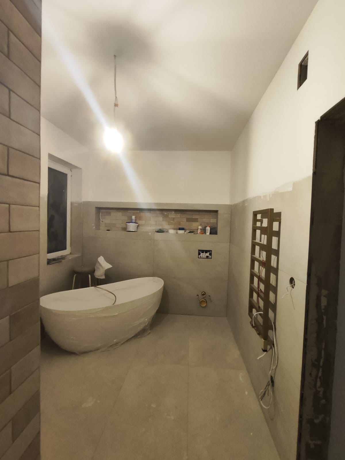 Łazienka w trakcie remontu: wolnostojąca wanna owinięta folią ochronną, ceramiczna zabudowa wnęki, grzejnik drabinkowy bez podłączenia, widoczne instalacje hydrauliczne i elektryczne.