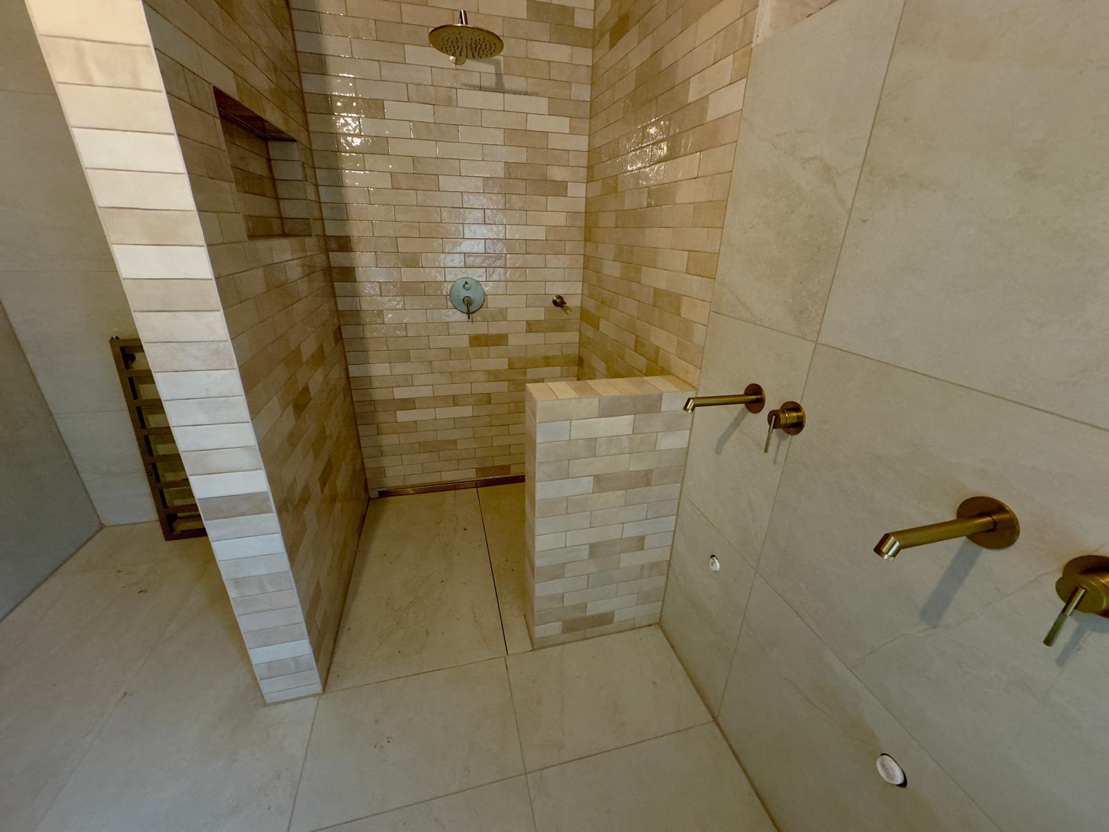 Wykończony prysznic walk-in z beżowymi płytkami imitującymi cegłę, złotą armaturą i odpływem liniowym w podłodze.
