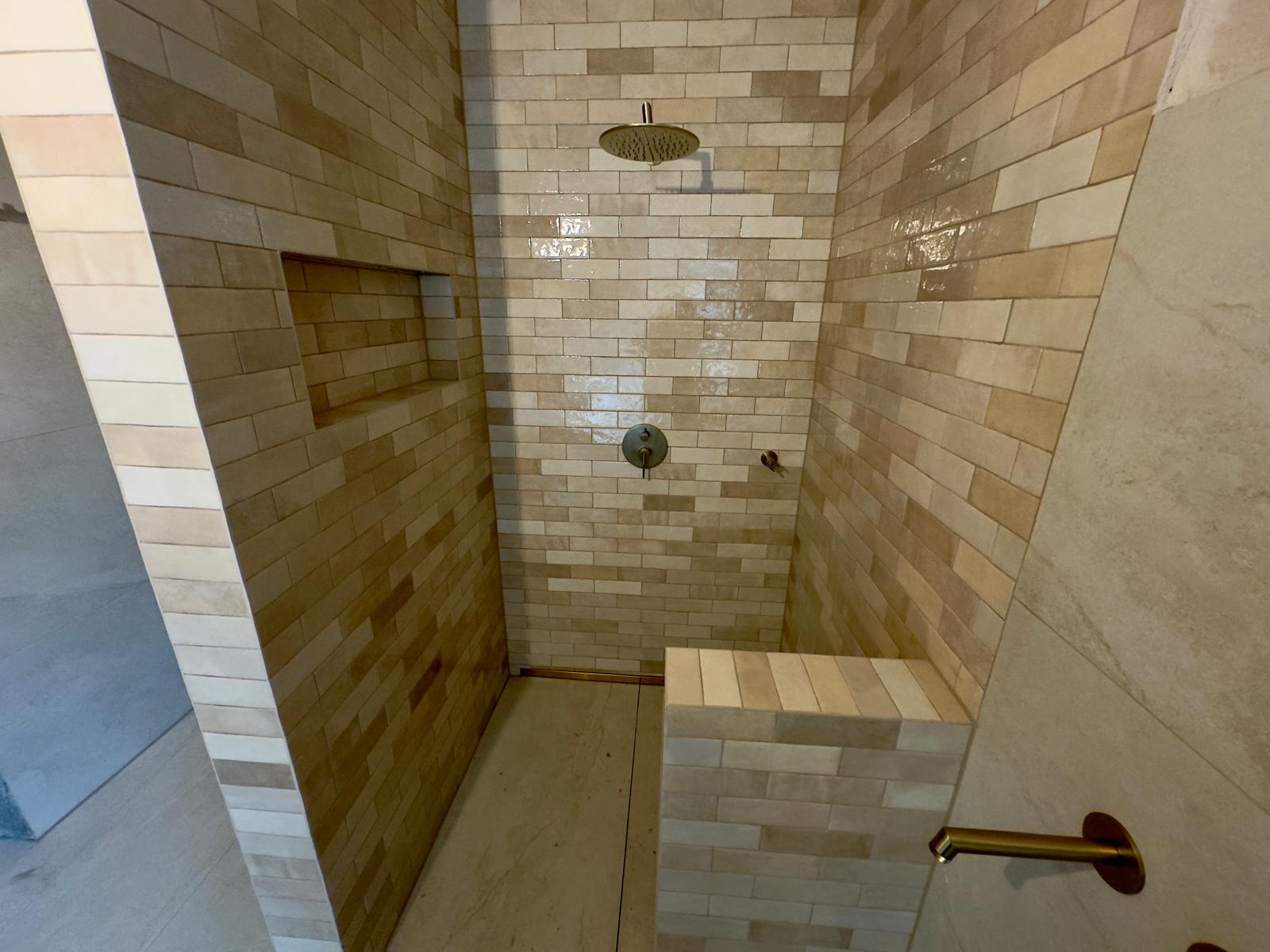 Wykończona kabina prysznicowa z beżowymi, prostokątnymi płytkami ułożonymi w cegiełkę, z wnęką w ścianie i siedziskiem, wyposażona w deszczownicę i baterię w kolorze mosiądzu.