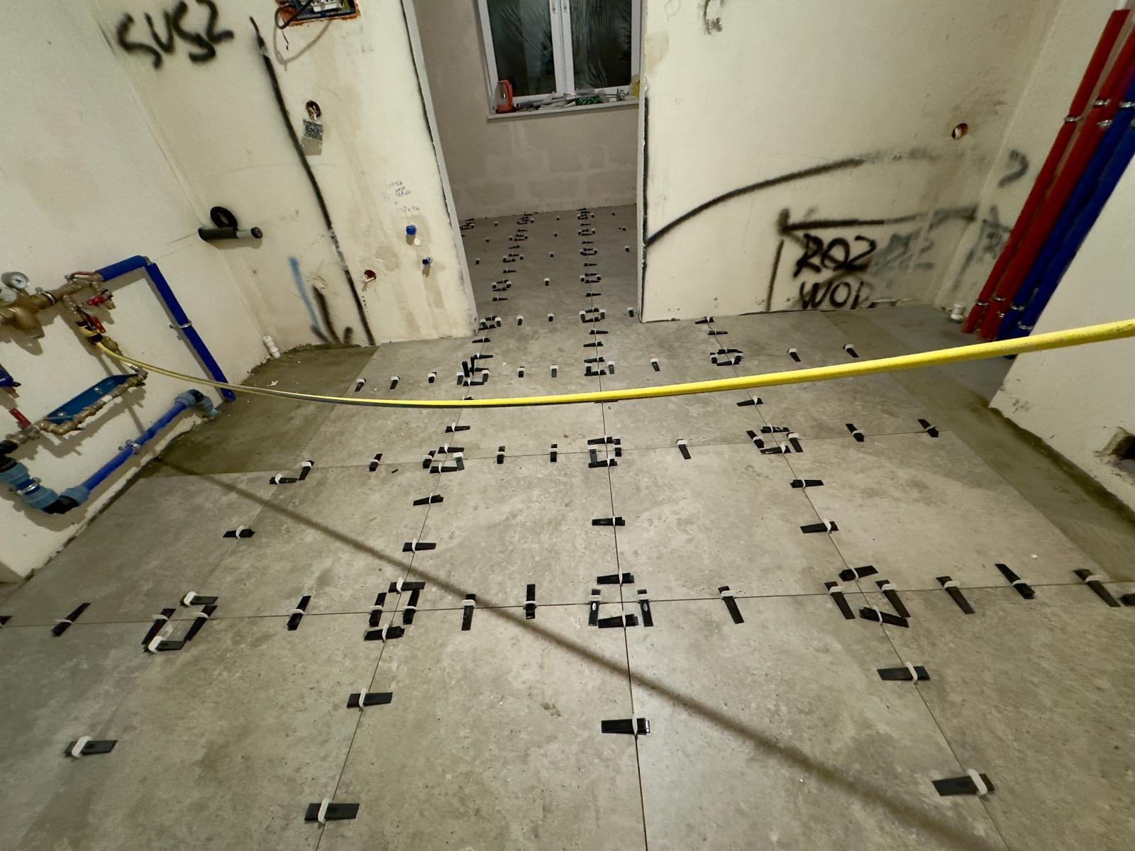Przygotowanie do ułożenia płytek podłogowych w łazience, widoczne krzyżyki dystansowe, rury instalacji wodnej i fragmenty graffiti na ścianach.