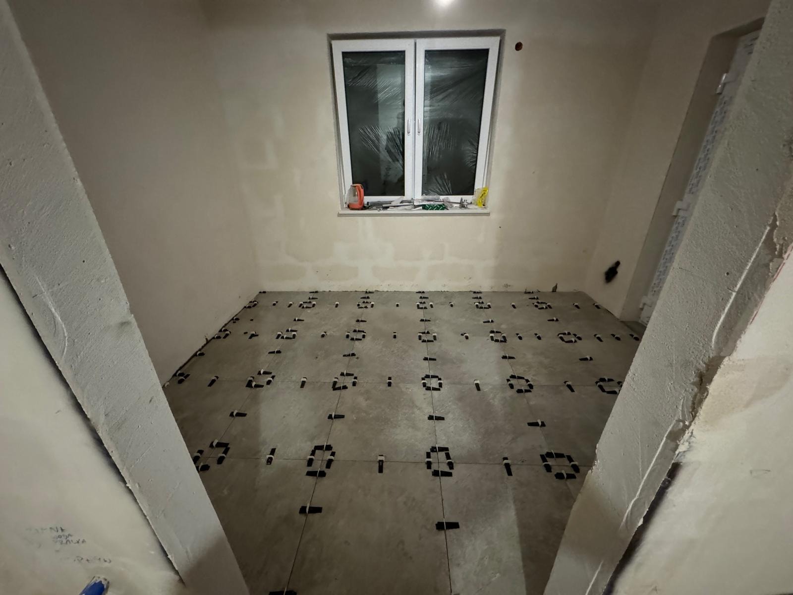 Proces układania szarych płytek podłogowych w pomieszczeniu z oknem, z użyciem systemu poziomowania płytek (klipsy i kliny), widoczne surowe ściany przygotowane do dalszych prac wykończeniowych.