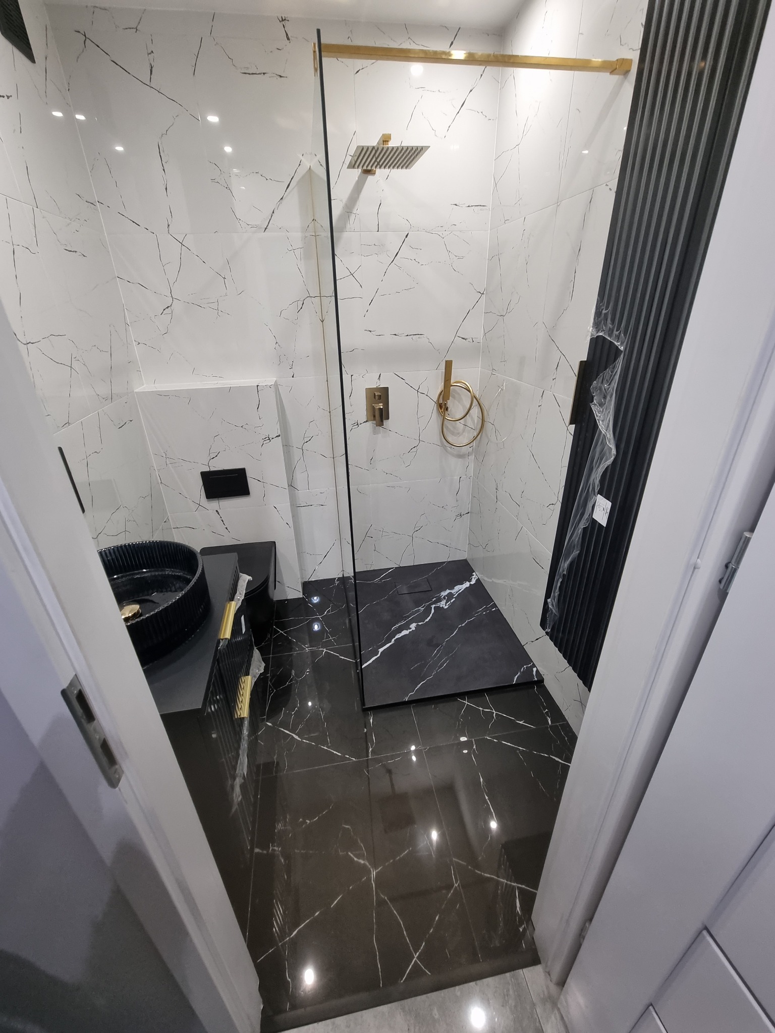 Nowoczesna łazienka z czarno-białymi płytkami imitującymi marmur, kabina prysznicowa typu walk-in z złotymi elementami, czarna umywalka nablatowa z ryflowanym wykończeniem, czarna armatura...