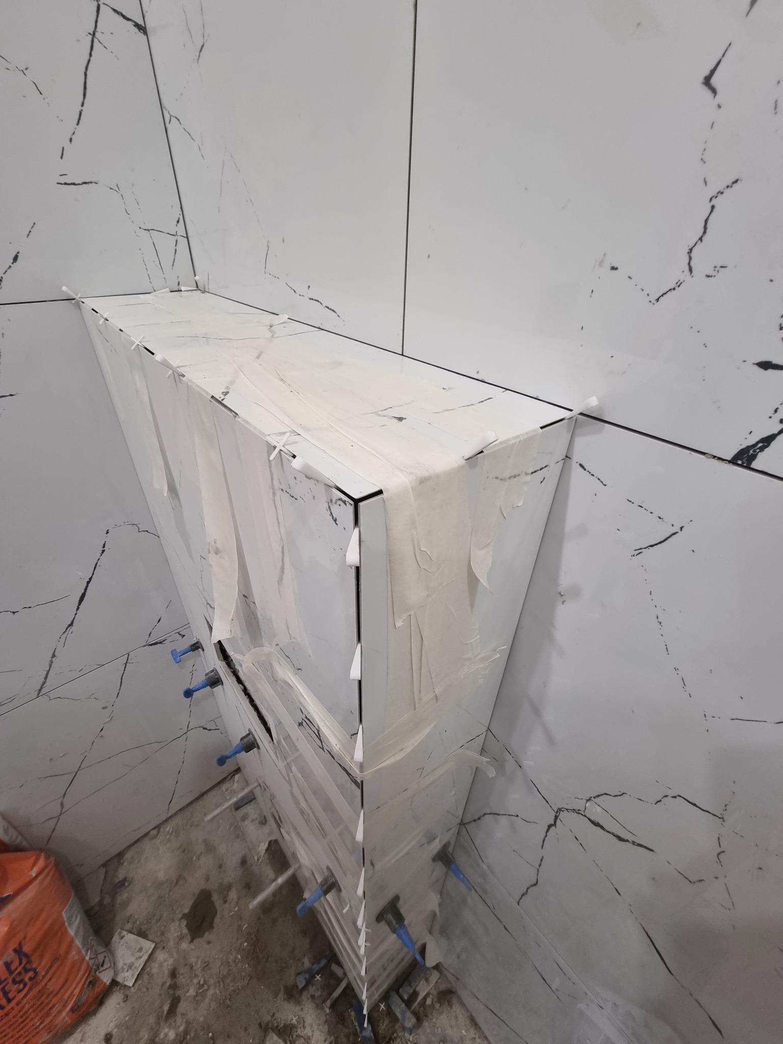 Półka w trakcie tworzenia z białych płytek imitujących marmur, zabezpieczona taśmą malarską i poziomowana niebieskimi klinami dystansowymi.