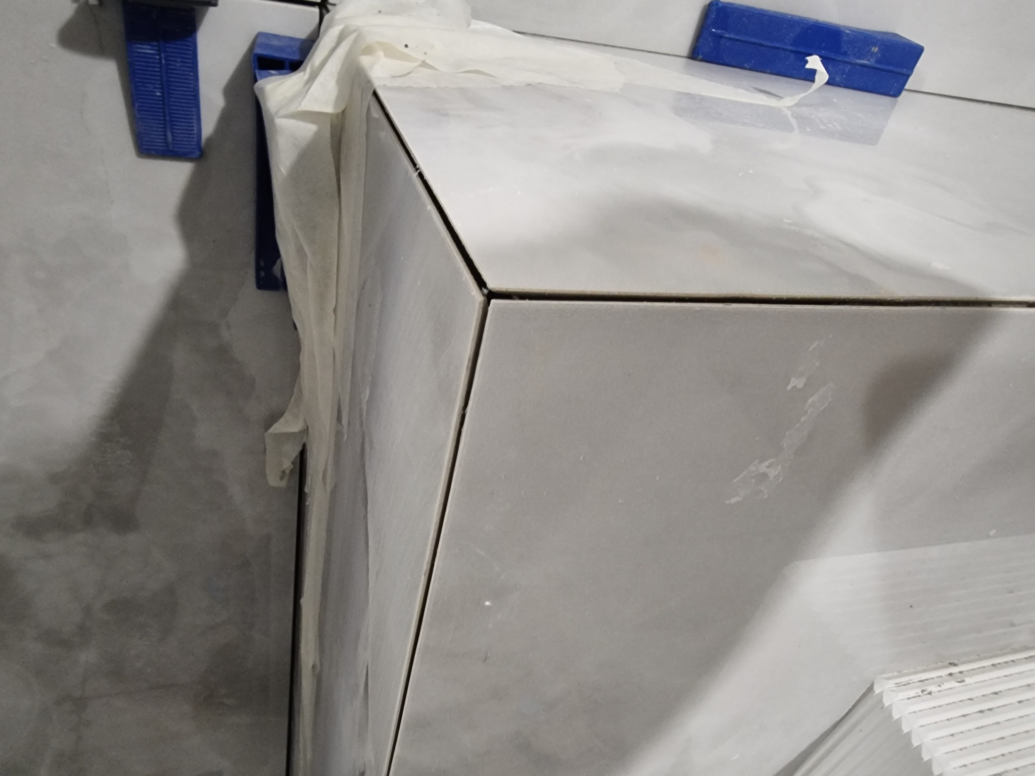 Fragment narożnika obłożonego jasnymi płytkami marmuropodobnymi, zabezpieczonego folią malarską i niebieskimi zaciskami, widoczne fugi i zapas płytek.