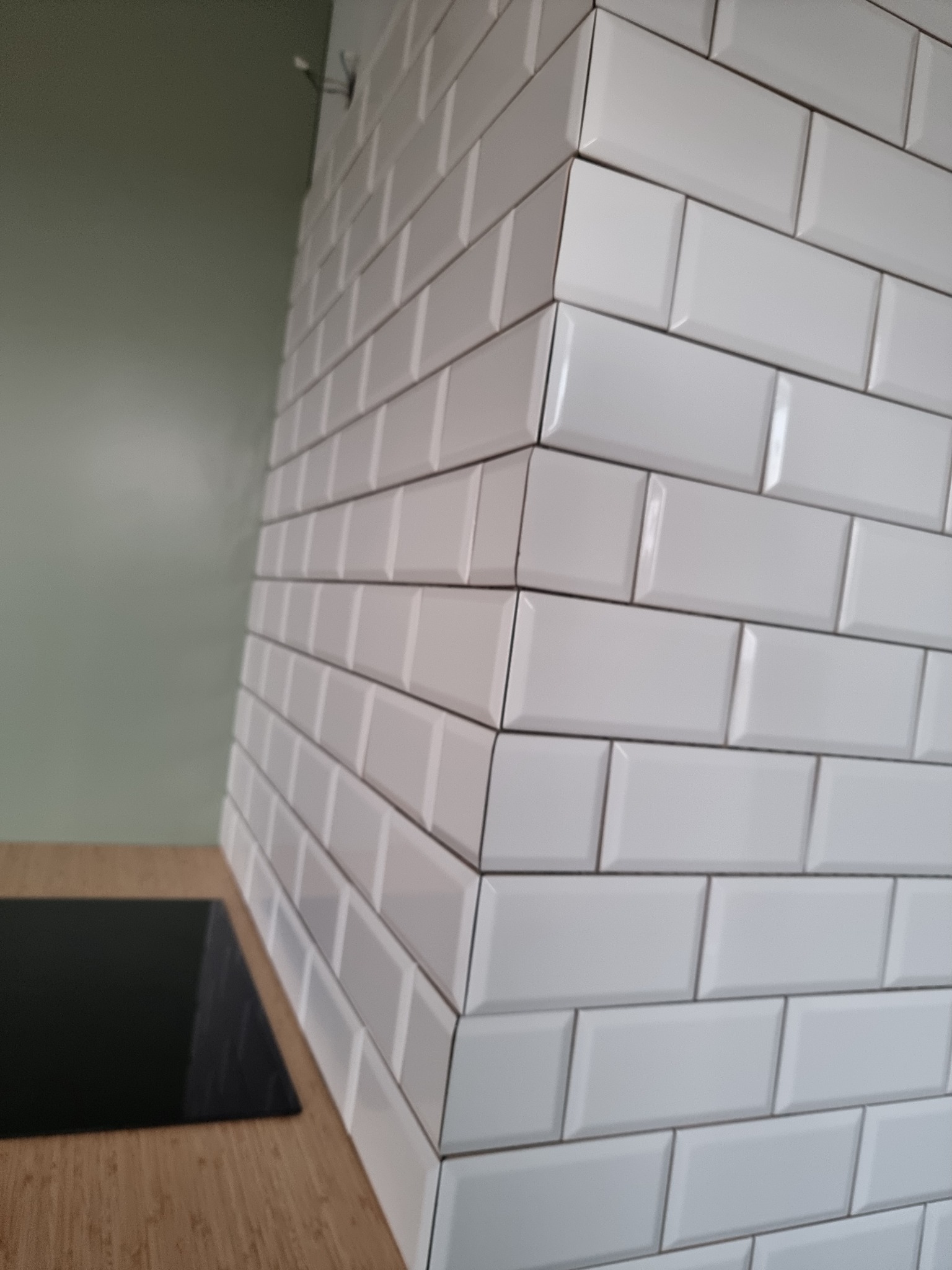 Białe, błyszczące płytki typu metro ułożone na ścianie nad blatem kuchennym z czarną płytą grzewczą, widoczne fugi w kolorze szarym, ściana pomalowana na zielono w tle.