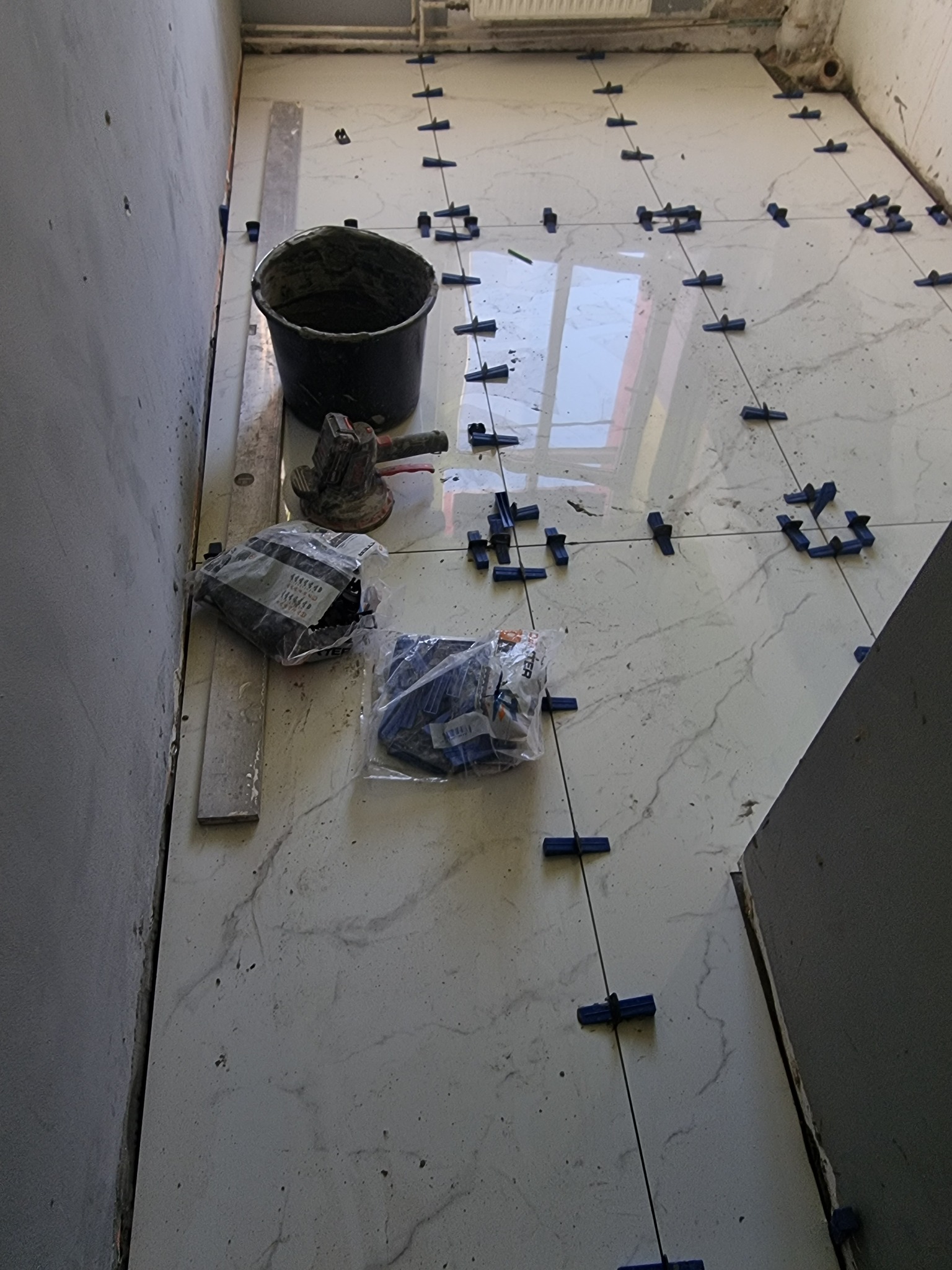 Białe płytki podłogowe z szarymi żyłkami układane z użyciem systemu poziomowania, widoczne narzędzia i wiadro z zaprawą.