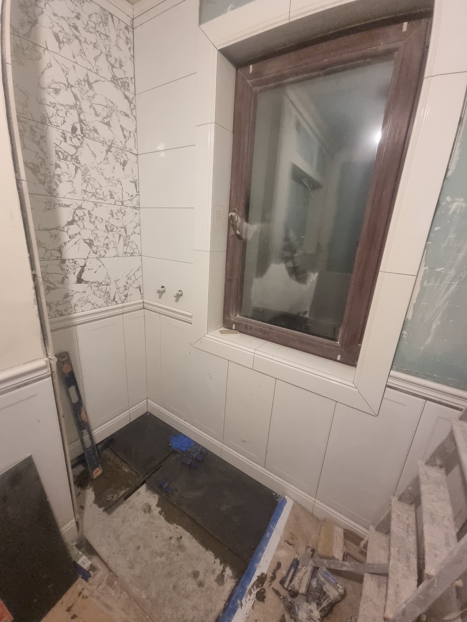 Łazienka w trakcie remontu: białe płytki ścienne, częściowo wykończone marmurowe panele, brązowe okno z białym obramowaniem, ciemne płytki podłogowe w trakcie układania, poziomica przy ścianie.