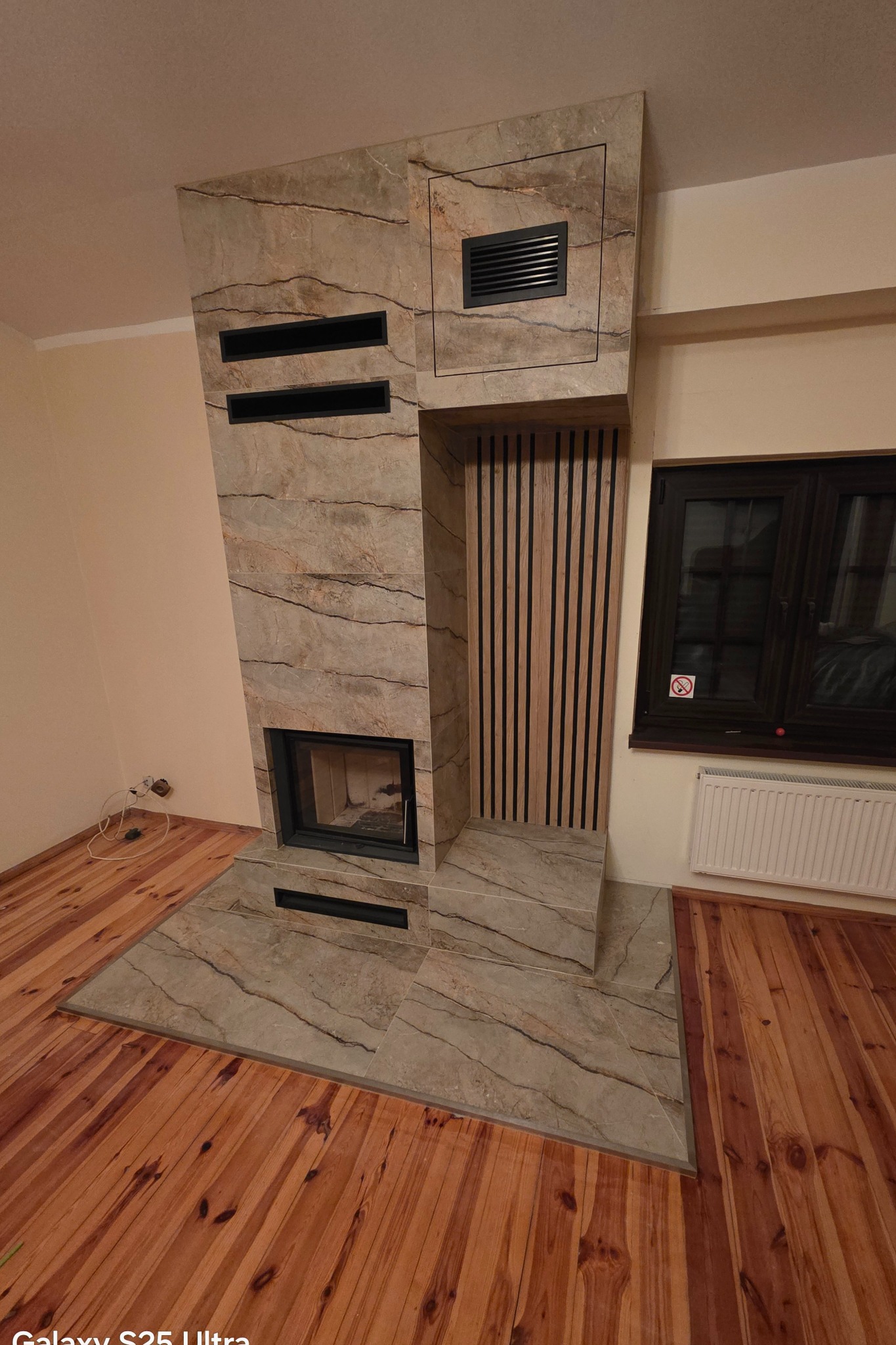 Wykończony kominek z płytkami imitującymi marmur, z drewnianymi panelami dekoracyjnymi. Widoczne detale wykończenia i połączenie materiałów.