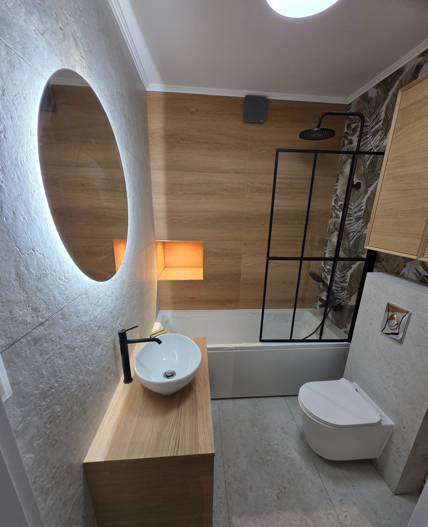 Nowoczesna łazienka z owalnym lustrem LED, drewnianą szafką pod umywalką i kabiną prysznicową z czarnymi ramami. Jasne płytki imitujące drewno i szaro-beżowa kolorystyka.