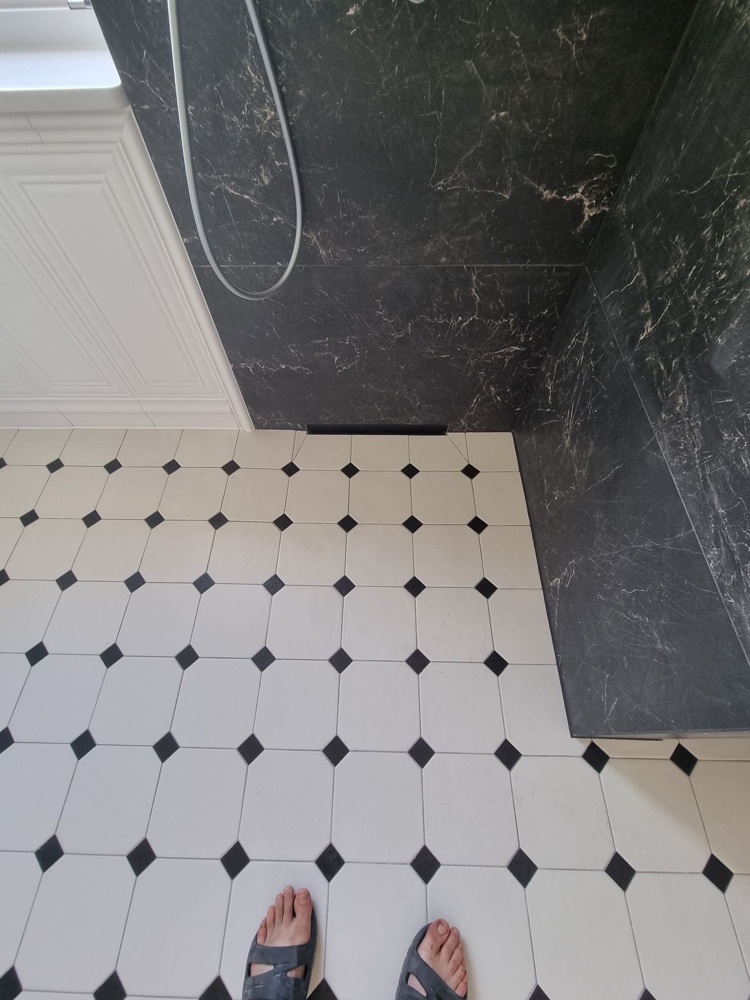 Wykończona łazienka z czarno-białą podłogą w karo, czarnymi marmurkowymi płytkami na ścianie prysznica i fragmentem białej, dekoracyjnej listwy przysufitowej.