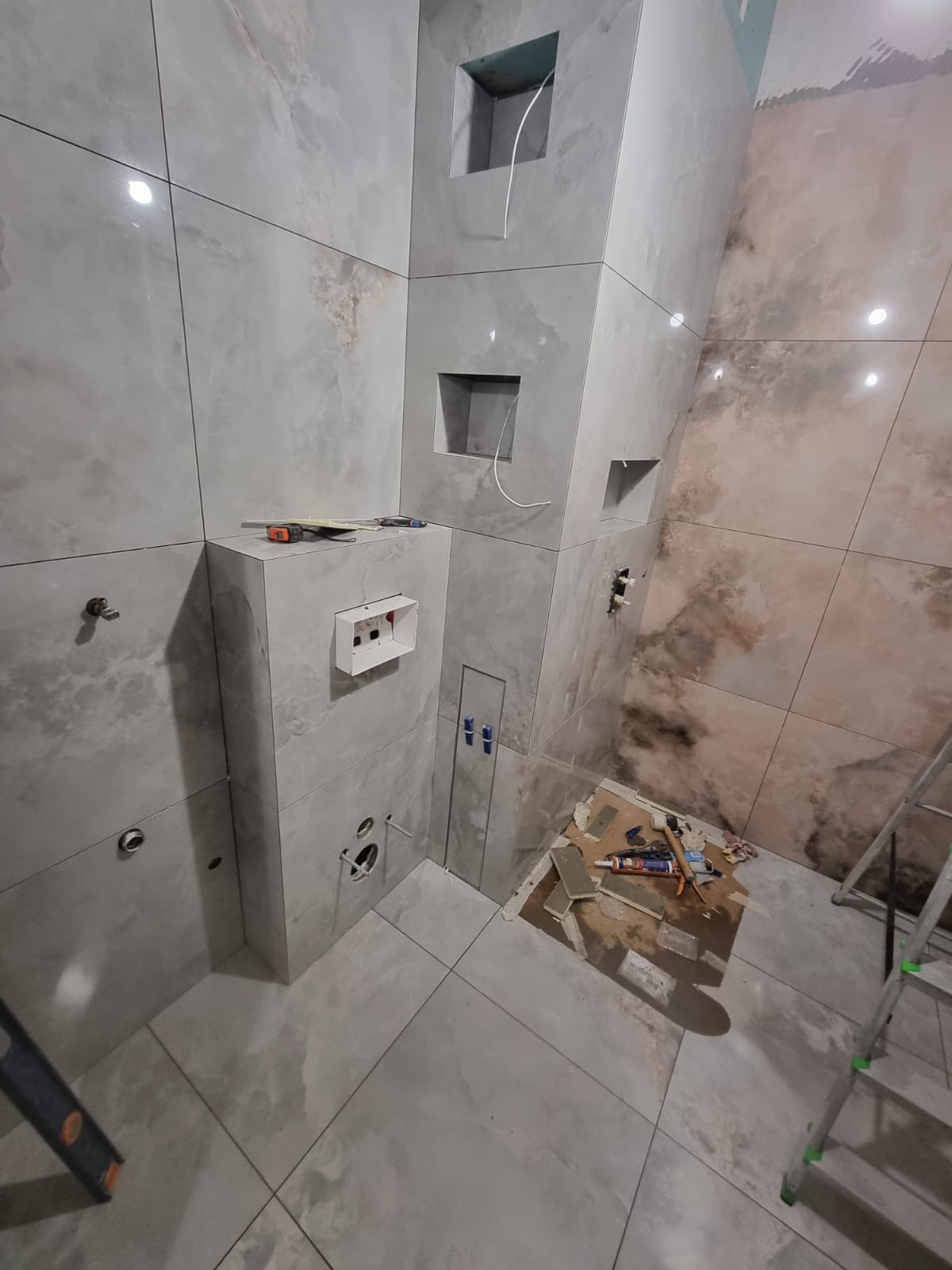 Kafelkowanie łazienki w trakcie: szare płytki imitujące kamień, widoczne narzędzia, otwory na armaturę, wnęki w ścianie. Częściowo ukończona realizacja.