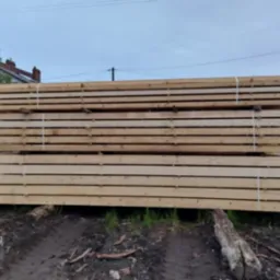 Stos świeżo przetartego drewna budowlanego, zabezpieczonego białym sznurkiem, ułożony na ziemi z widocznymi śladami opon, w tle drzewa i fragment domu.