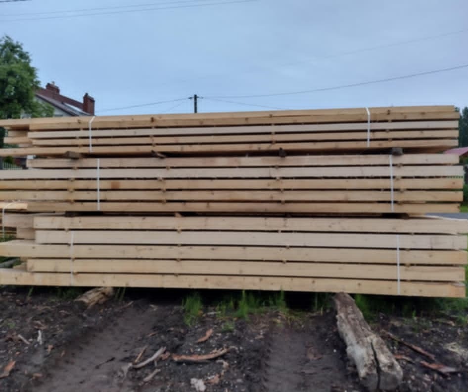 Stos świeżo przetartego drewna budowlanego, zabezpieczonego białym sznurkiem, ułożony na ziemi z widocznymi śladami opon, w tle drzewa i fragment domu.