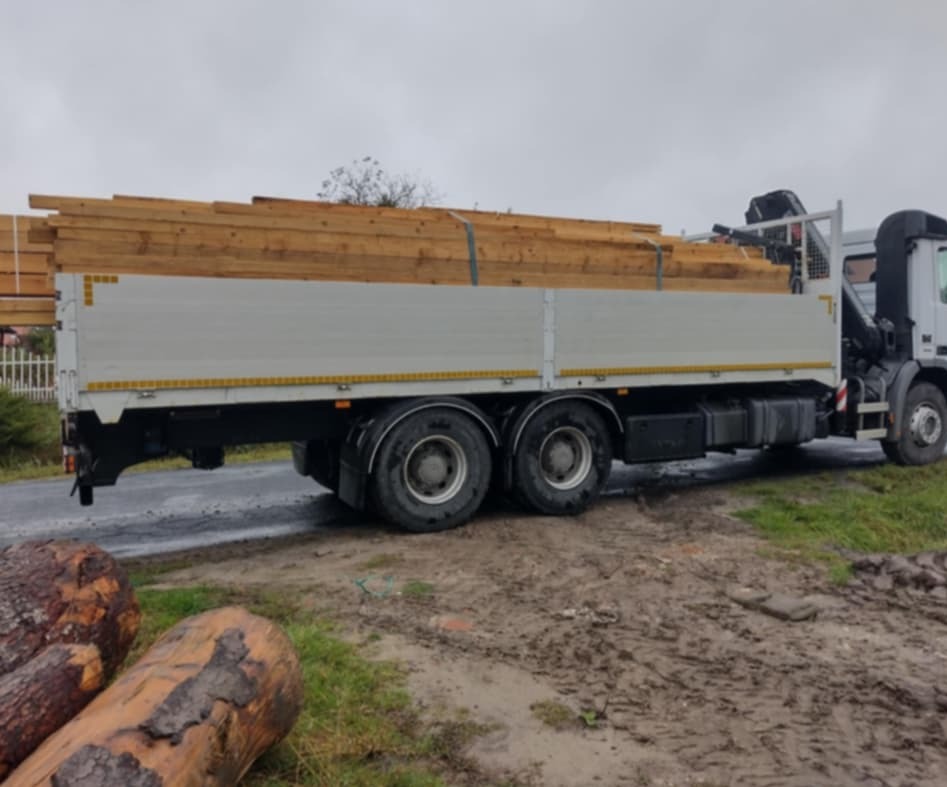 Ciężarówka z platformą załadowana drewnem budowlanym, zaparkowana na błotnistej drodze, widoczne ścięte pnie drzew