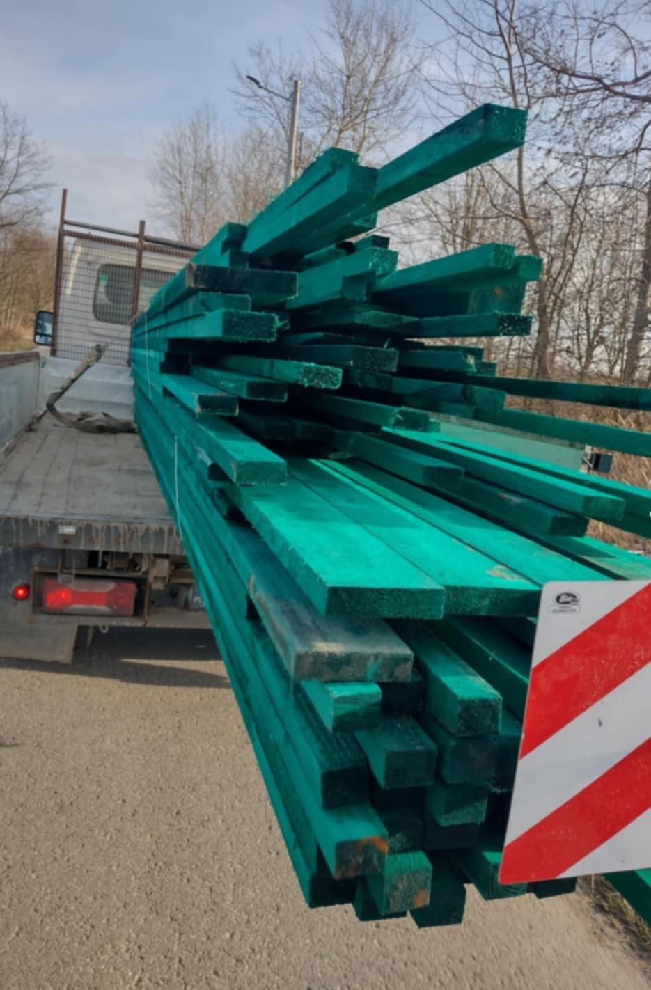 Transport belek zaimpregnowanego na zielono drewna na pace ciężarówki, widoczny znak ostrzegawczy.