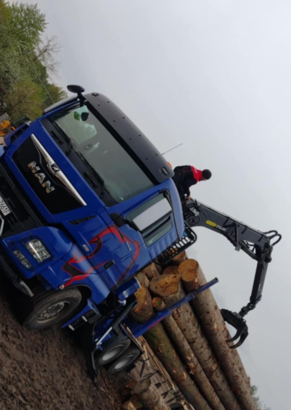 Niebieska ciężarówka MAN z hydraulicznym ramieniem załadowczym, przewożąca pnie drzew. Operator w czerwonej kurtce kontroluje załadunek. Widoczne błoto na kołach i ziemi.