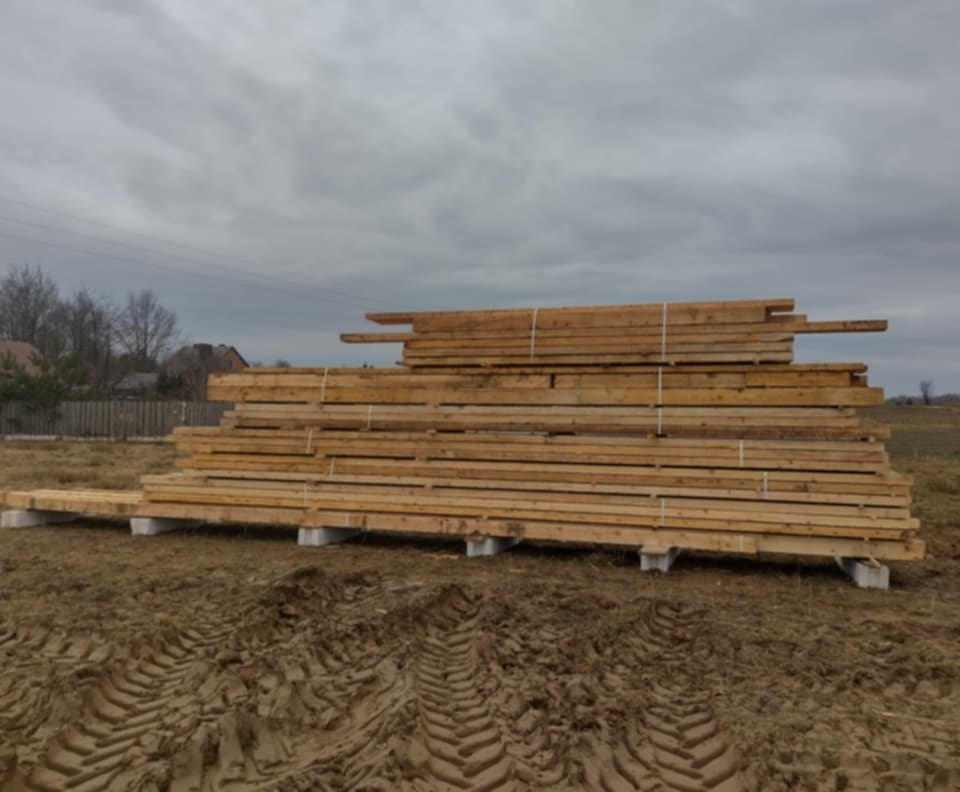 Stos desek na betonowych podporach, zabezpieczony białymi taśmami, na błotnistym terenie w Żołyni, pochmurne niebo w tle.