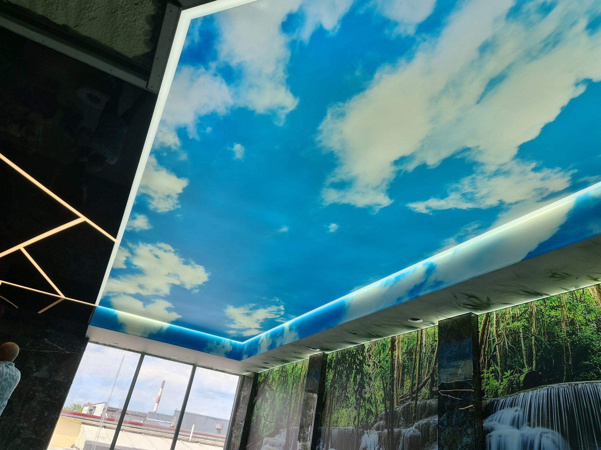 Jasny, podświetlany sufit z motywem nieba i chmur, zainstalowany w Słupsku, z widocznymi elementami dekoracyjnymi na ścianach przedstawiającymi motyw lasu i wodospadu.