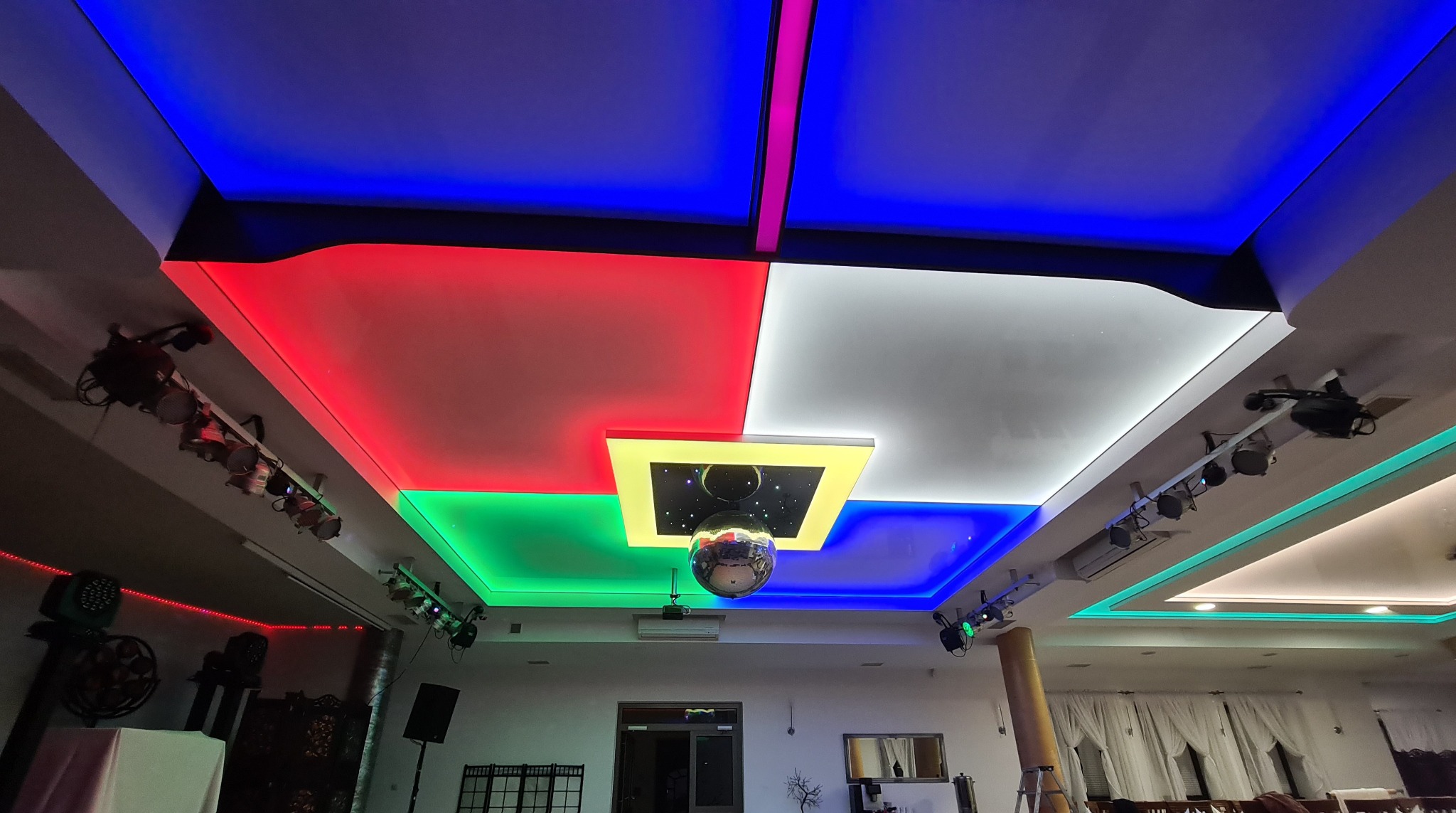 Wielobarwny sufit napinany z podświetleniem LED w kolorach niebieskim, czerwonym, zielonym i białym, z centralnie umieszczoną kulą dyskotekową i kwadratową oprawą oświetleniową z motywem kosmicznym.