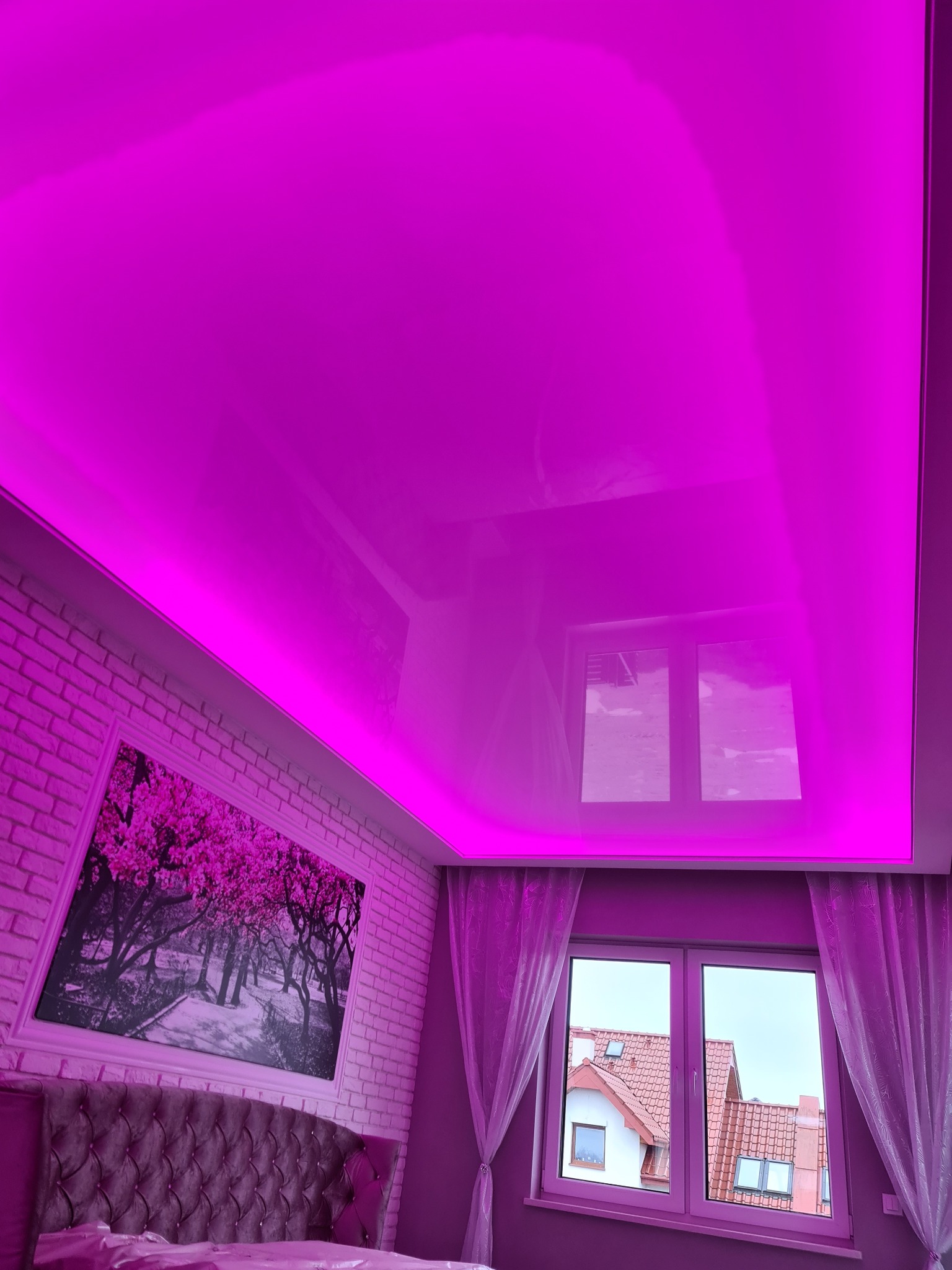 Błyszczący, fuksjowy sufit z oświetleniem LED wzdłuż krawędzi, odbijający widok z okna i obraz na ścianie z cegły nad wezgłowiem łóżka.