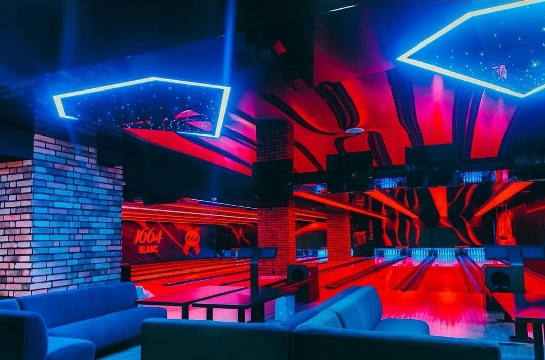Wnętrze kręgielni z podwieszonymi sufitami z motywem gwiazd i neonowym oświetleniem, ceglana ściana z logo piwa 1664 Blanc, sofy w kolorze niebieskim.