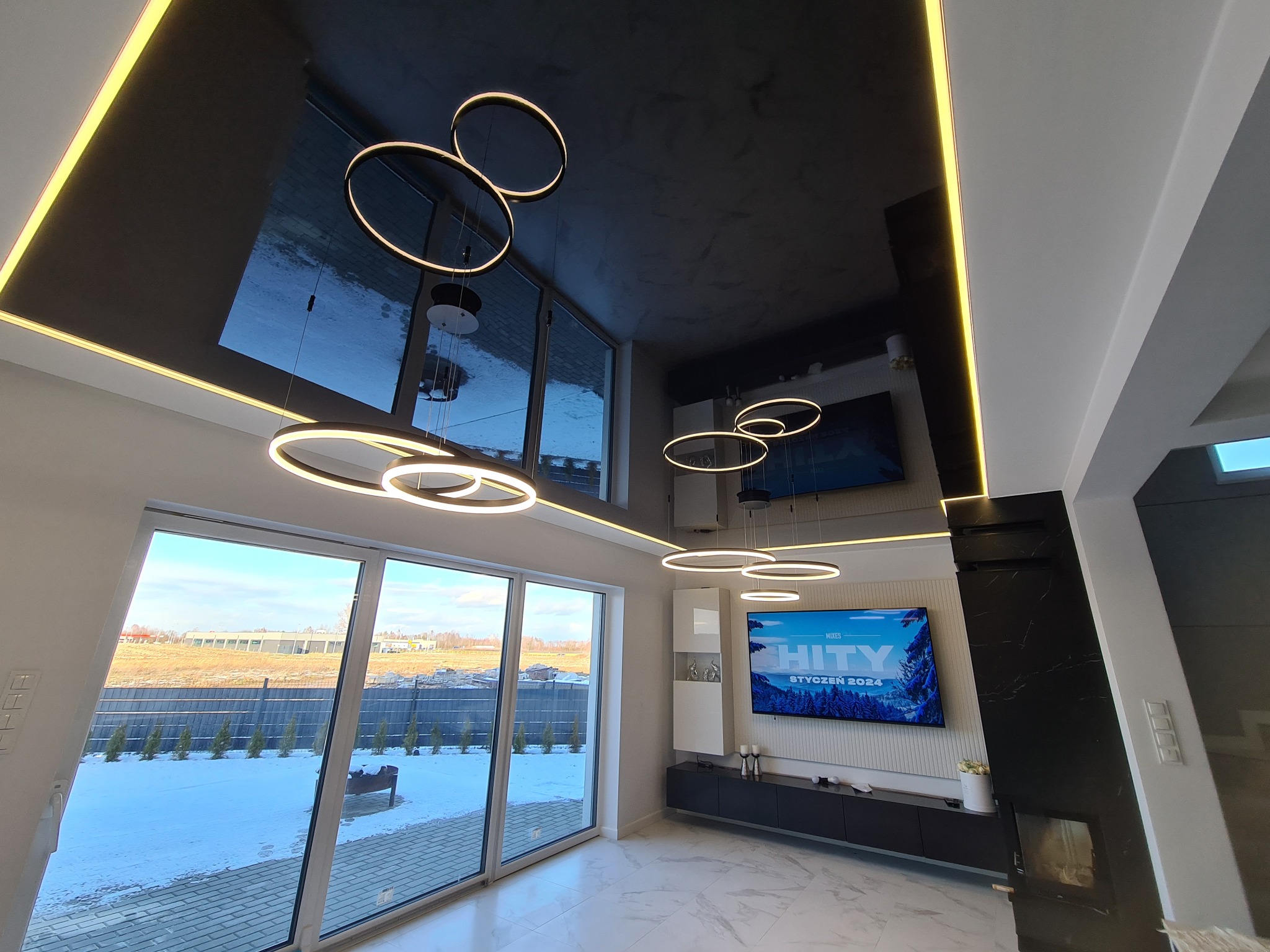Nowoczesny salon z czarnym, lustrzanym sufitem, oświetlony okrągłymi lampami LED, widok na zaśnieżony ogród przez panoramiczne okno.