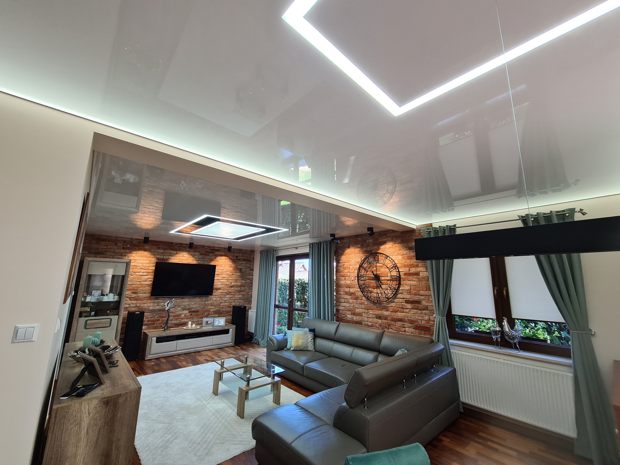 Błyszczący, biały sufit napinany z wbudowanym oświetleniem LED w kształcie kwadratów, odbijający wnętrze salonu z ceglaną ścianą, szarą sofą i elementami dekoracyjnymi.