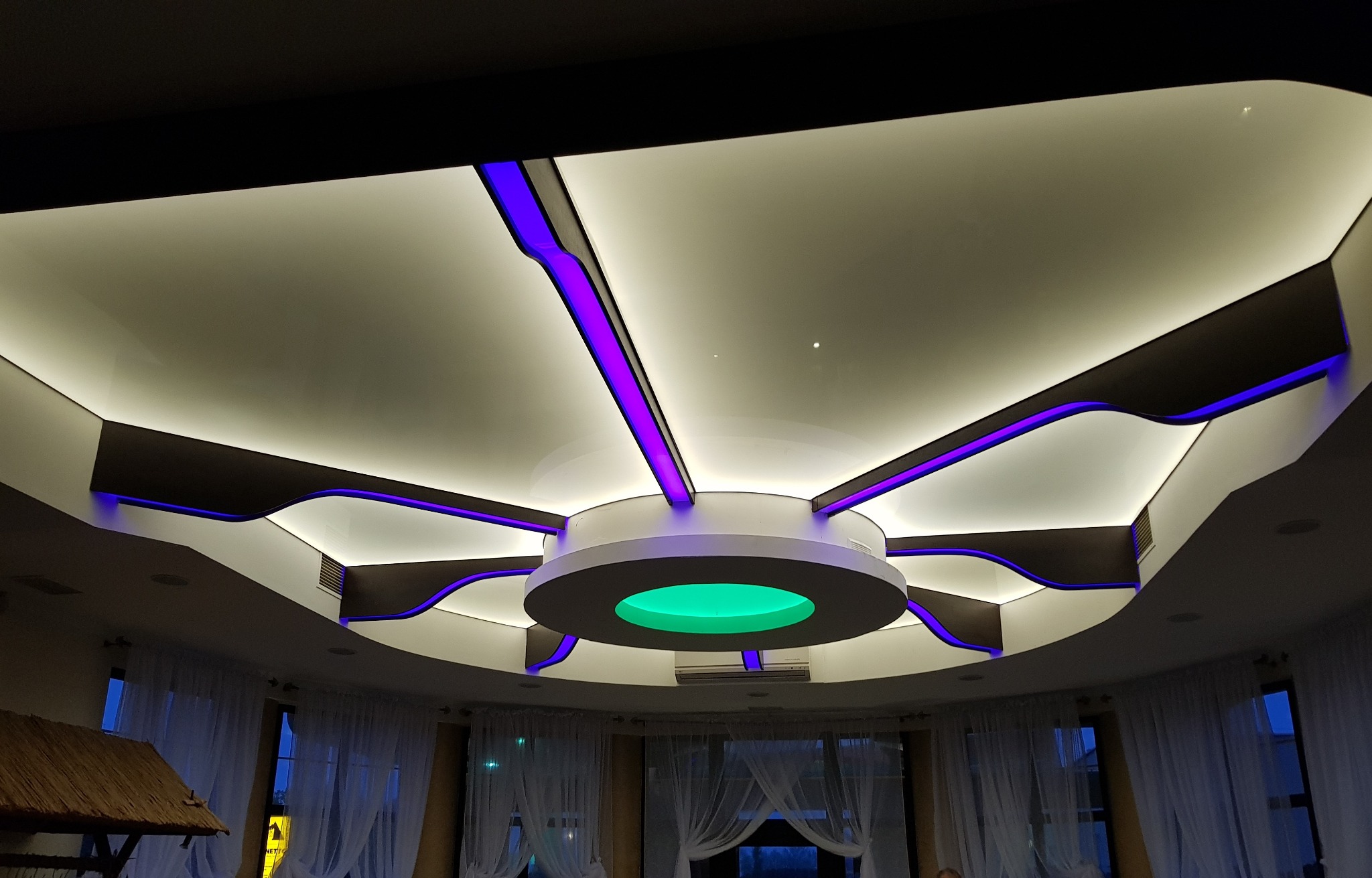 Nowoczesny sufit z podwieszanymi elementami i oświetleniem LED w kolorze fioletowym i zielonym, widok z dołu, w tle okna z białymi firankami.