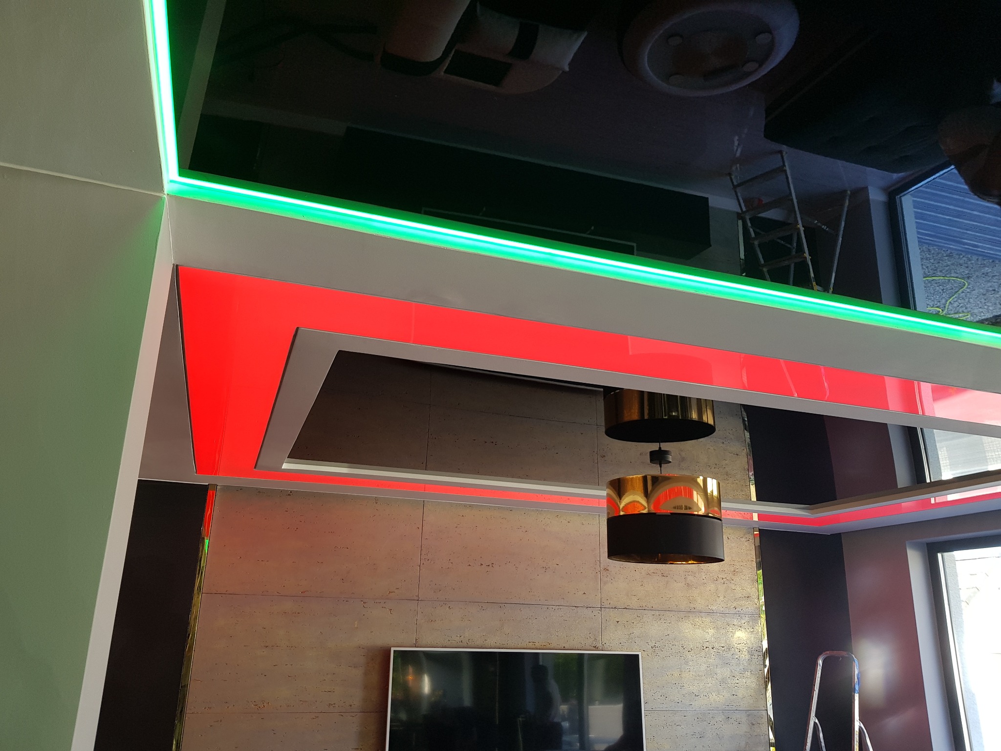 Błyszczący, czarny sufit z podświetleniem LED w kolorach czerwonym i zielonym, odbijający wnętrze pokoju z lampą i drabiną.