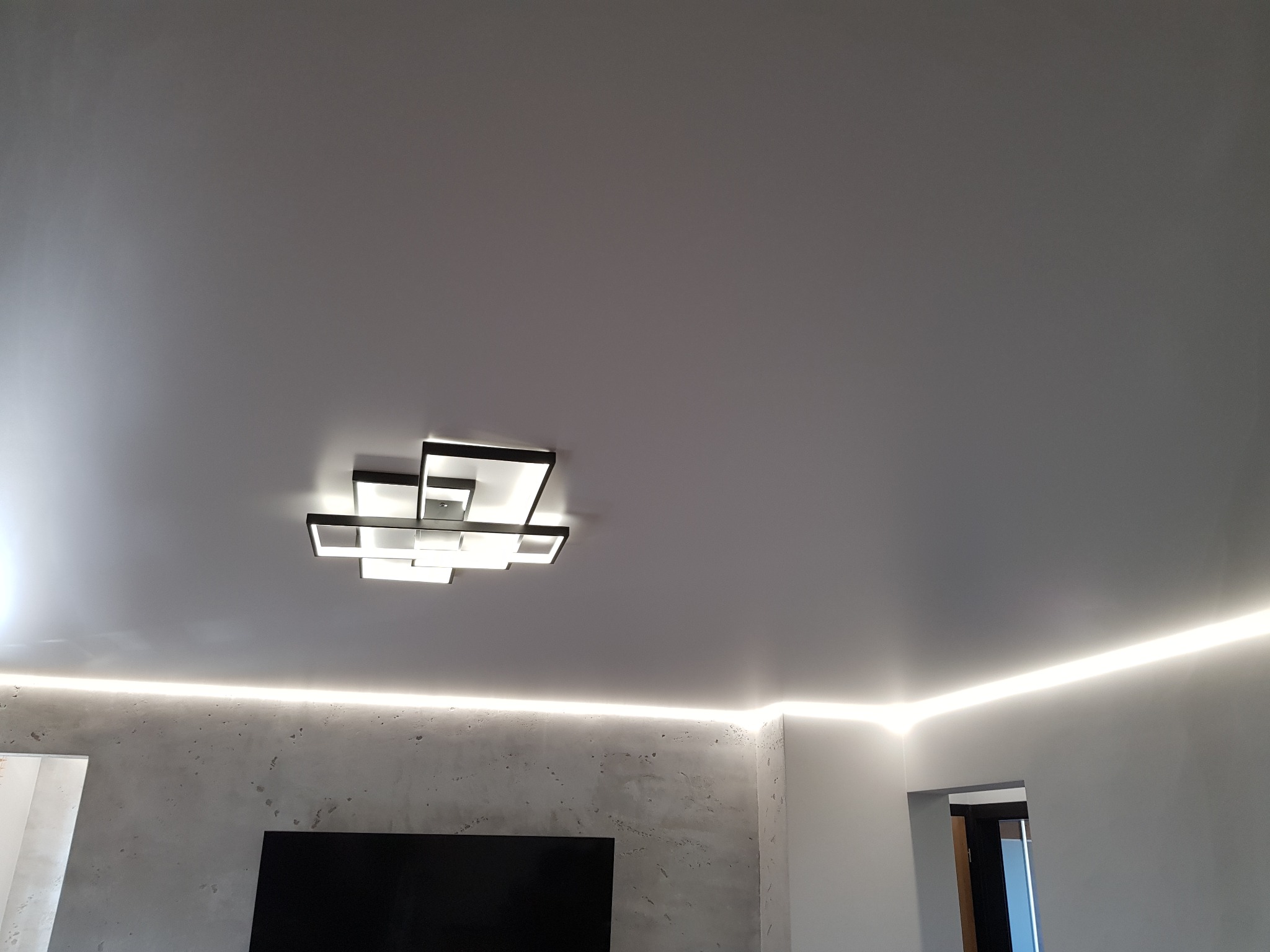 Biały, matowy sufit z podświetleniem LED wzdłuż krawędzi i nowoczesną lampą sufitową złożoną z kwadratów.