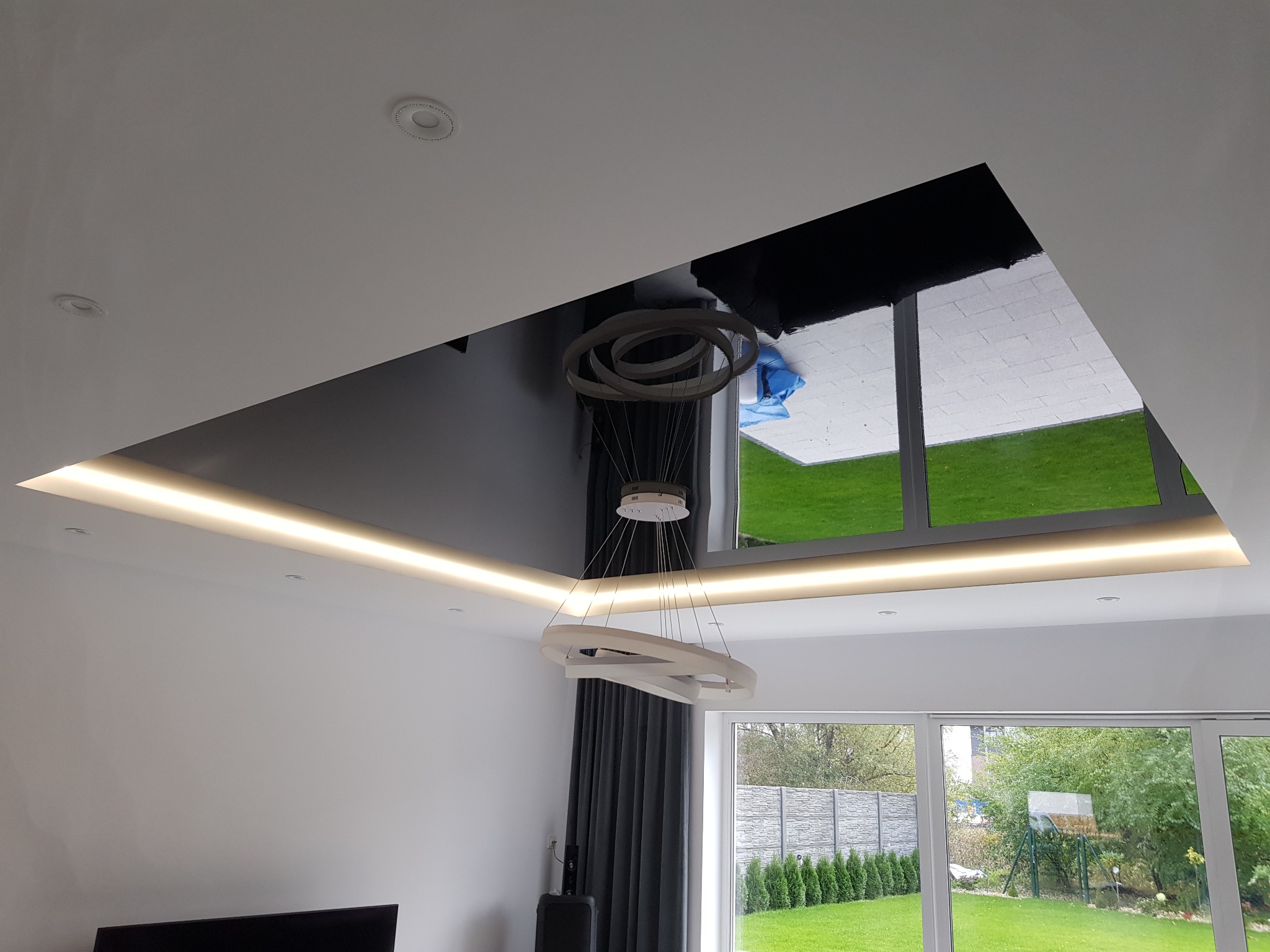 Nowoczesny salon z czarnym, błyszczącym wykończeniem sufitu, podświetleniem LED i designerską lampą wiszącą na tle okna z widokiem na ogród.
